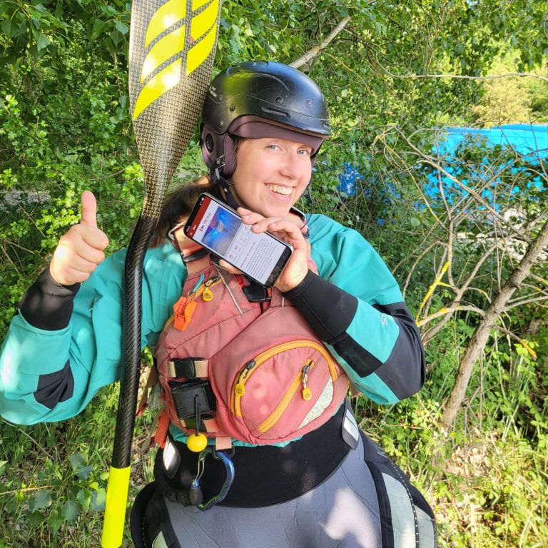 HKCW Vereinsapp Person hält ein Smartphone am Fluss, mit der HKCW-Vereinsapp-Anzeige auf dem Bildschirm.