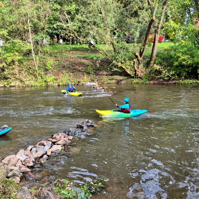 Glückliche Paddlerinnen beim HKCW Zwei Personen in Neoprenanzügen mit Schwimmwesten, lächelnd an einem Ufer.