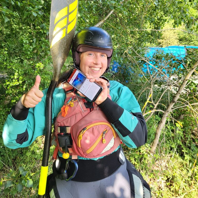HKCW Vereinsapp Person hält ein Smartphone am Fluss, mit der HKCW-Vereinsapp-Anzeige auf dem Bildschirm.